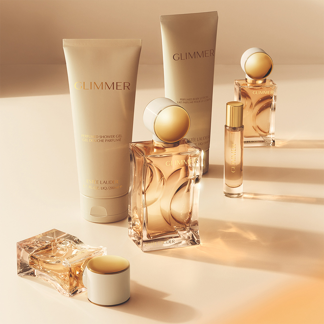 Estée Lauder Glimmer fragrance collection with golden perfume bottles, body lotion, and shower gel arranged on neutral surface