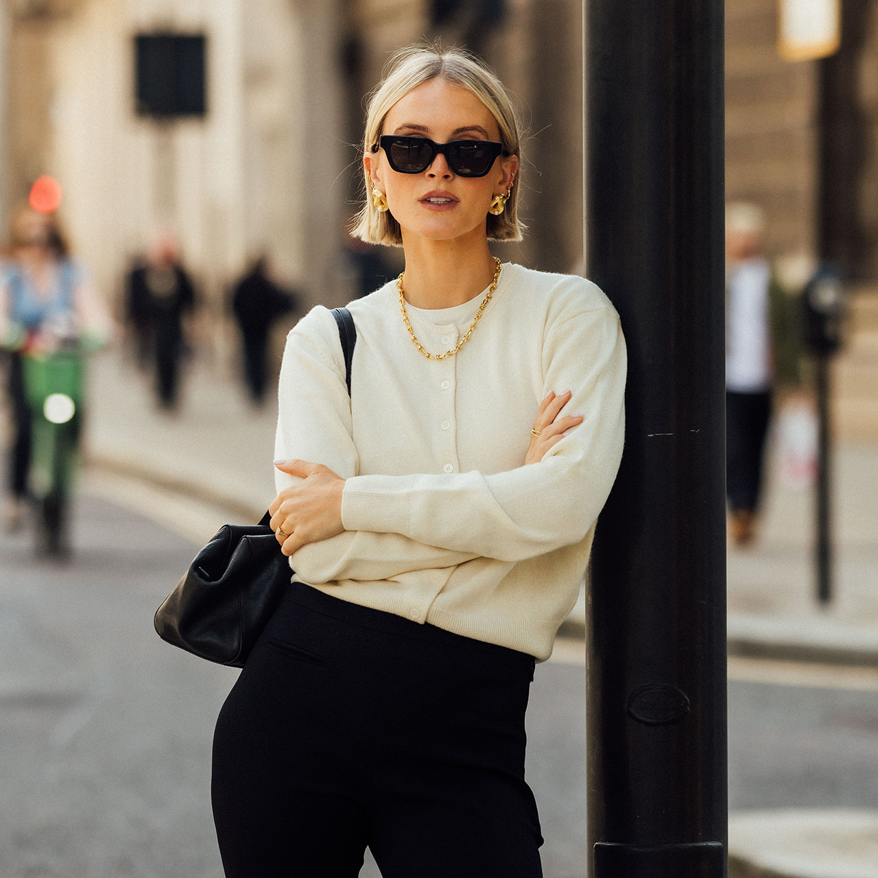 Woman on city street wearing cream cardigan, black trousers, gold chain necklace and sunglasses