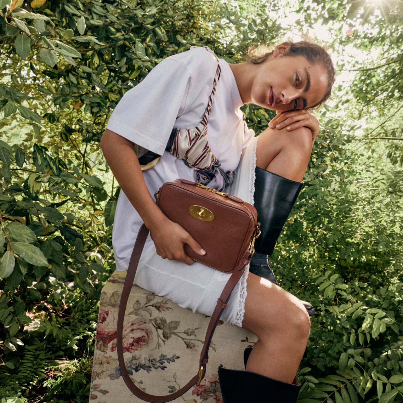 Woman in white shirt holding brown leather crossbody bag, surrounded by green foliage
