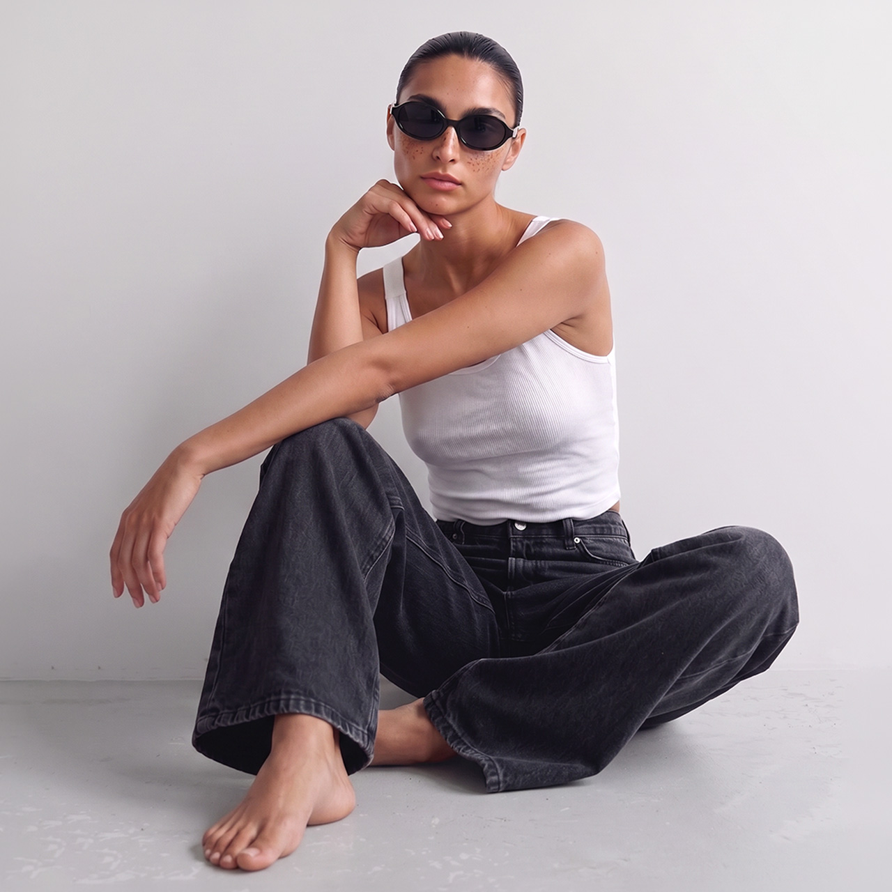 Woman wearing white sleeveless top and black denim jeans, sitting with sunglasses