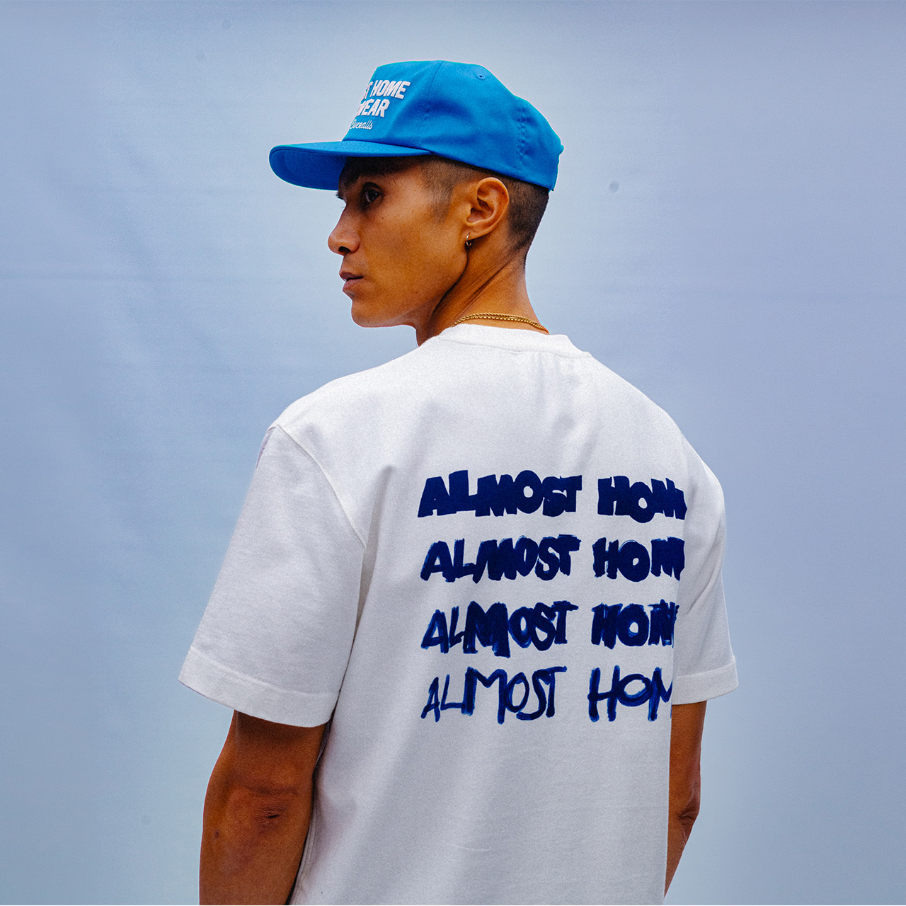 Man wearing white t-shirt with blue repeated text graphic and blue cap against clear blue sky backdrop