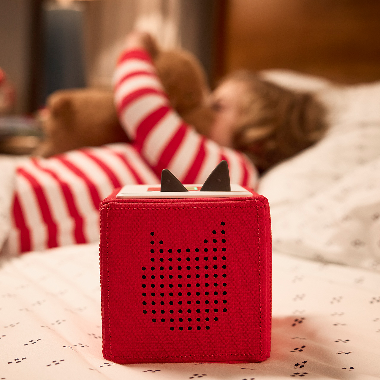 Red fabric Cuddle Tonie with cat ears and speaker grille, child wearing red and white striped socks in background