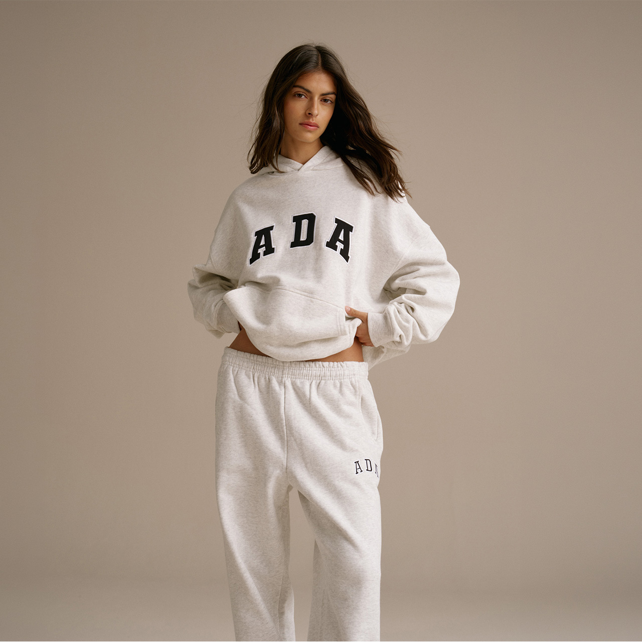 Woman wearing cream marl hoodie and matching joggers with ADA branding, standing against neutral background