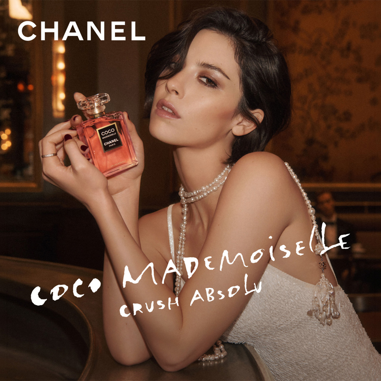 Woman holding red Coco Mademoiselle Crush Absolu fragrance bottle against gold-lit backdrop