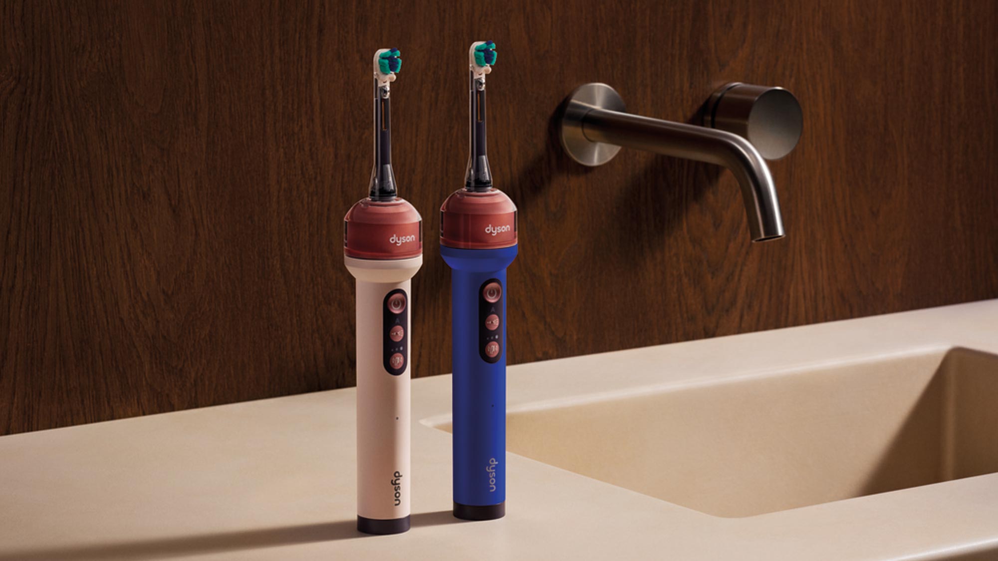 Two electric toothbrushes stand upright on a beige bathroom counter next to a modern faucet. One toothbrush is light pink, while the other is dark blue, both featuring red accents and multiple buttons. The background is a dark wooden wall.