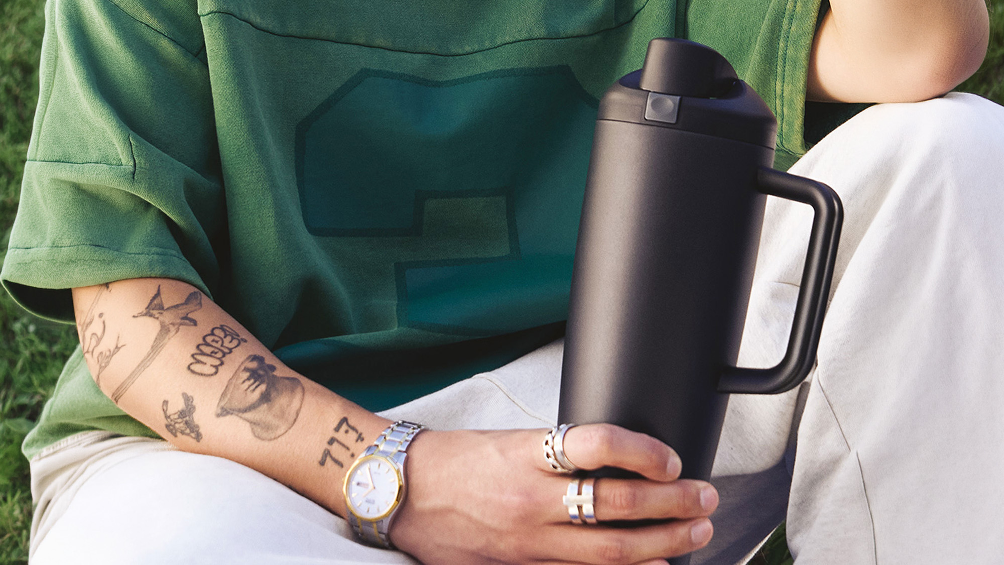 Person wearing green shirt and white trousers holding black insulated water bottle with handle, wearing watch and rings