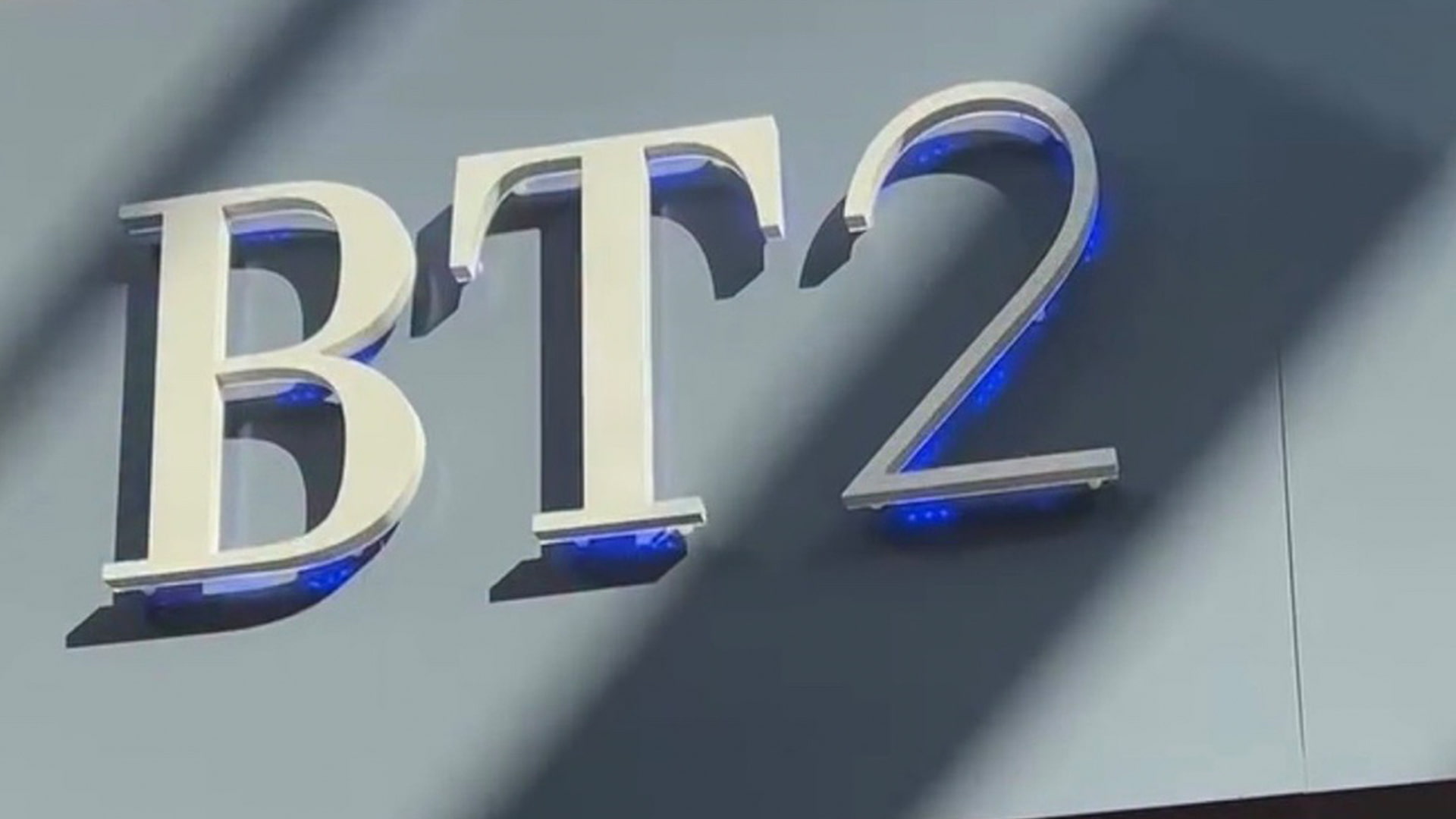 'BT2' in large white letters with blue neon accents on a blue-grey background. Sunlight casts shadows across the wall.