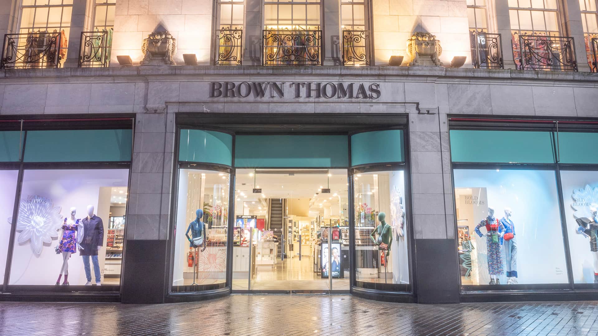 The image shows the exterior of Brown Thomas, an upscale department store. The building features large glass windows displaying elegantly dressed mannequins. Inside, the store is brightly lit, showcasing various luxury items. The pavement outside is wet, suggesting recent rain.
