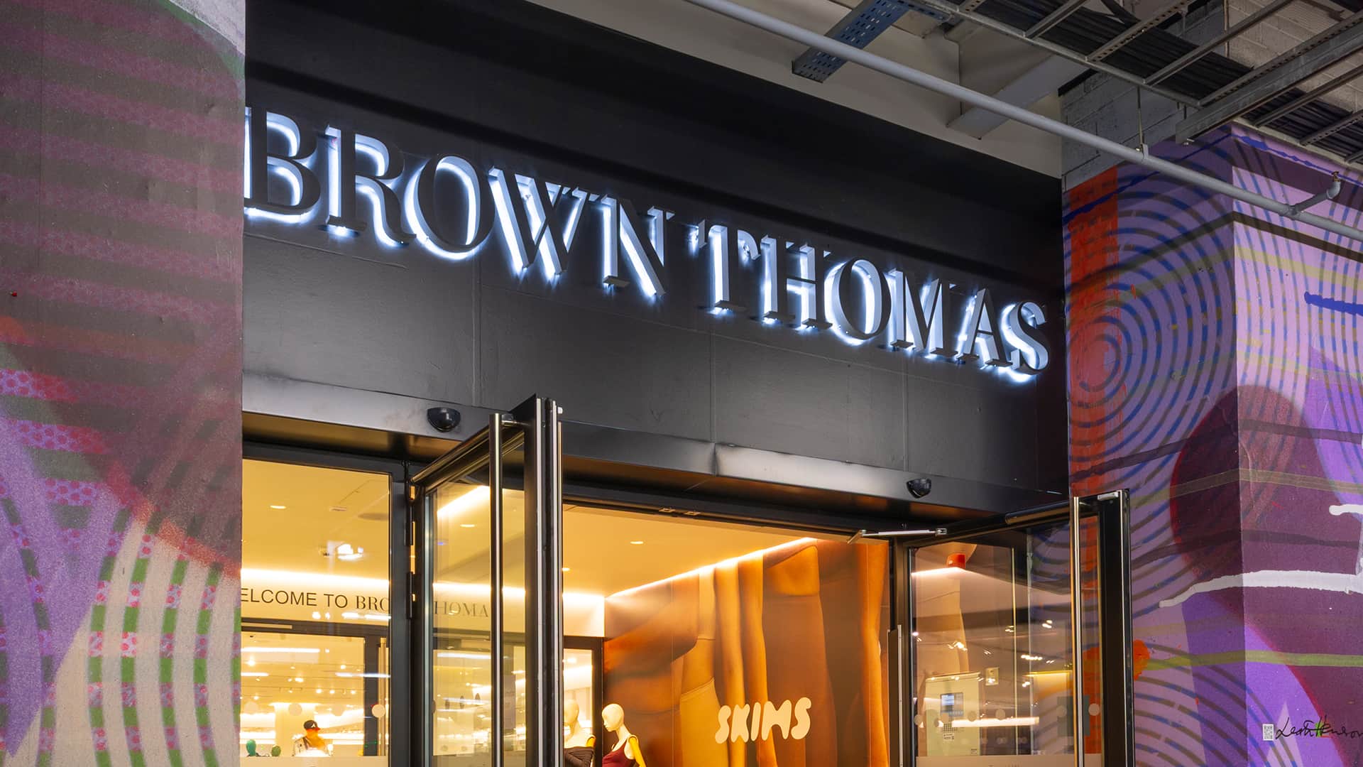 Entrance of a Brown Thomas store with illuminated signage. The interior is visible, showing a well-lit display area. Colourful abstract patterns decorate the exterior walls surrounding the entrance.