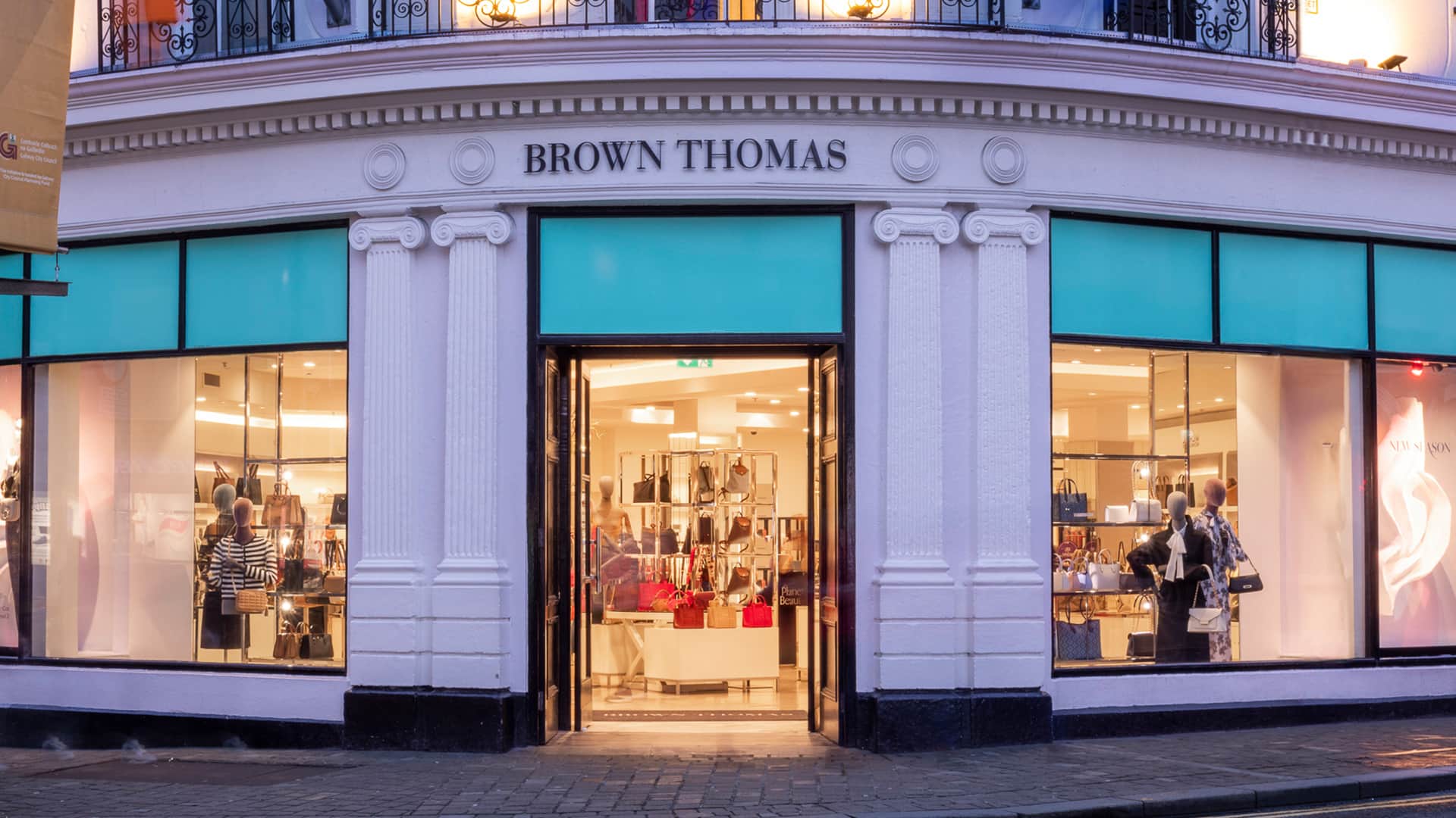 Entrance of the Brown Thomas department store, featuring large glass windows displaying various luxury items. The exterior is elegantly designed with decorative columns and bright, inviting lighting. Shoppers are visible through the windows.
