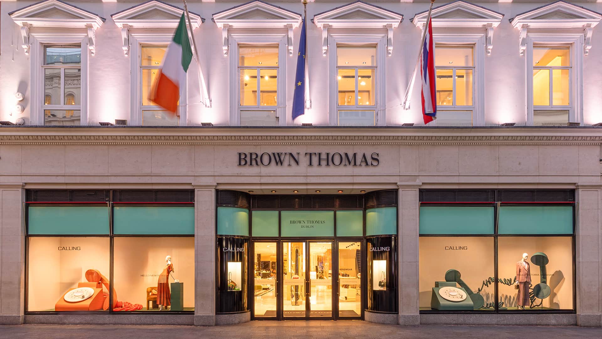 The front of Brown Thomas department store is illuminated at night. Three national flags, including the Irish flag, are displayed above. Large windows reveal elegantly arranged displays, and the entrance is centrally positioned, creating a welcoming appearance.