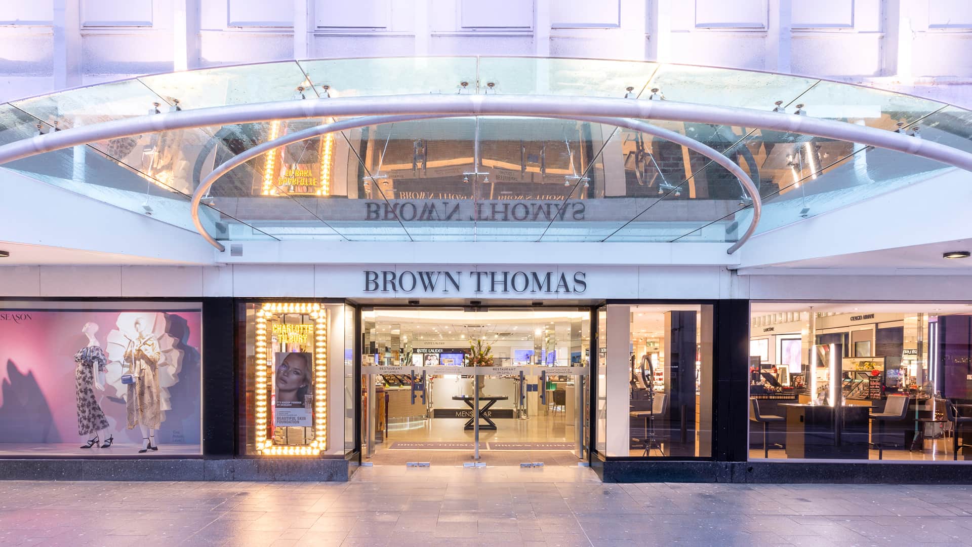 The entrance of Brown Thomas, a department store, featuring large glass doors flanked by display windows with mannequins in elegant clothing. Above the entrance, a glass canopy with the store's name in bold letters adds a modern touch.