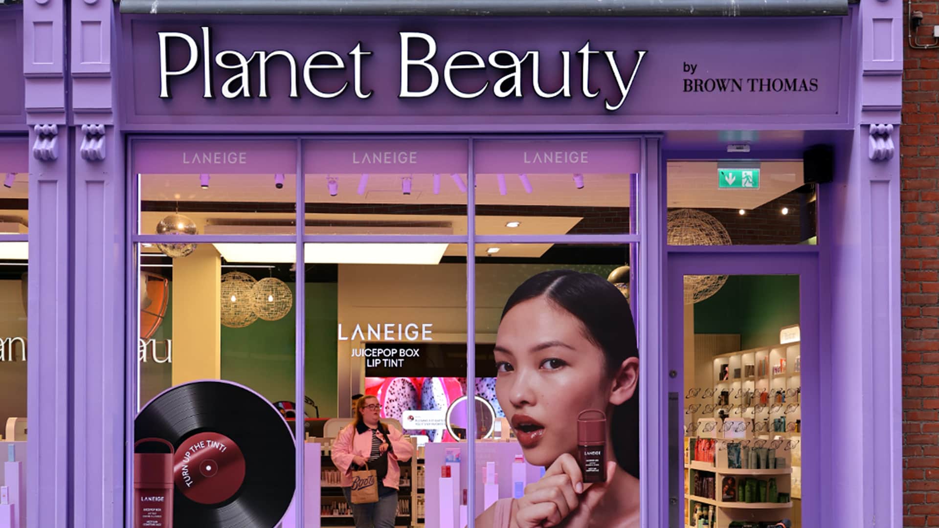 Purple storefront of Planet Beauty by Brown Thomas, featuring a large poster of a woman holding a cosmetic product. Interior displays are visible through the window, showcasing beauty items.