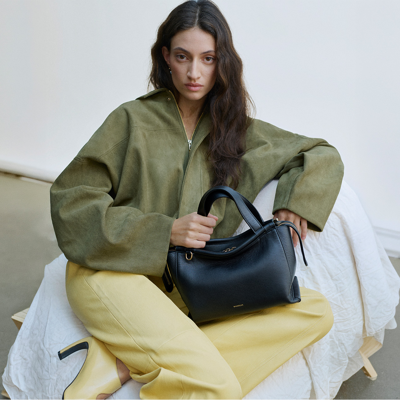 Woman wearing olive green jacket and yellow trousers, holding black leather handbag