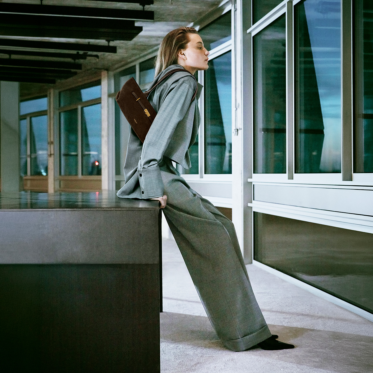 Woman in grey blazer carrying brown leather shoulder bag in modern office corridor with green-tinted windows