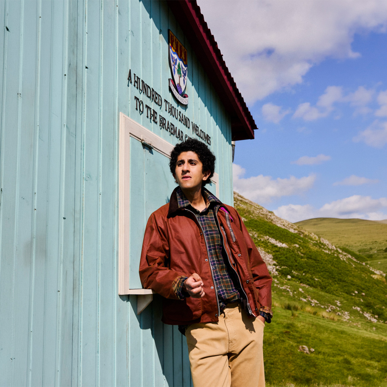 Man wearing rust-coloured jacket and checked shirt standing outside rural building with rolling green hills