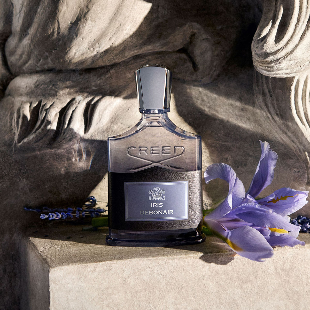 Creed Iris Debonair fragrance bottle with purple iris flowers and decorative beads on textured stone background