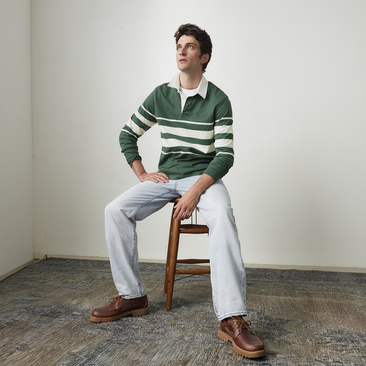 Man seated on wooden chair wearing green and white striped rugby shirt, light blue jeans, and brown shoes against neutral background
