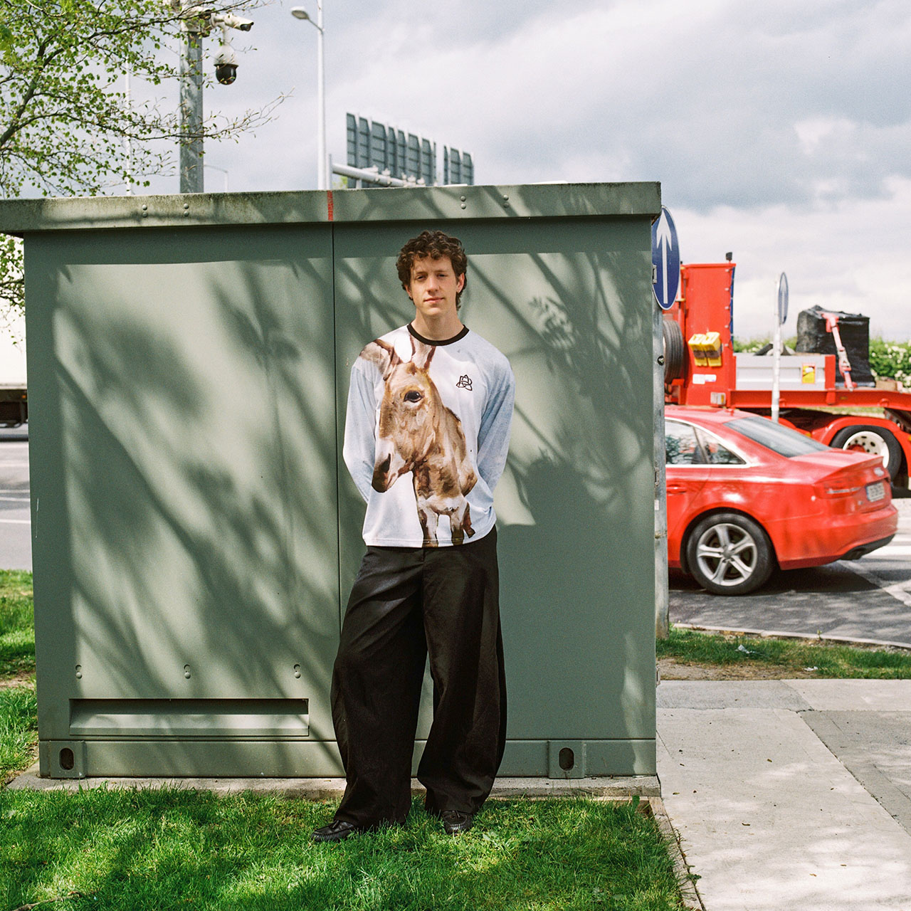 Young man holding a brown dog, wearing light blue long-sleeve shirt and black trousers, standing in front of green storage unit