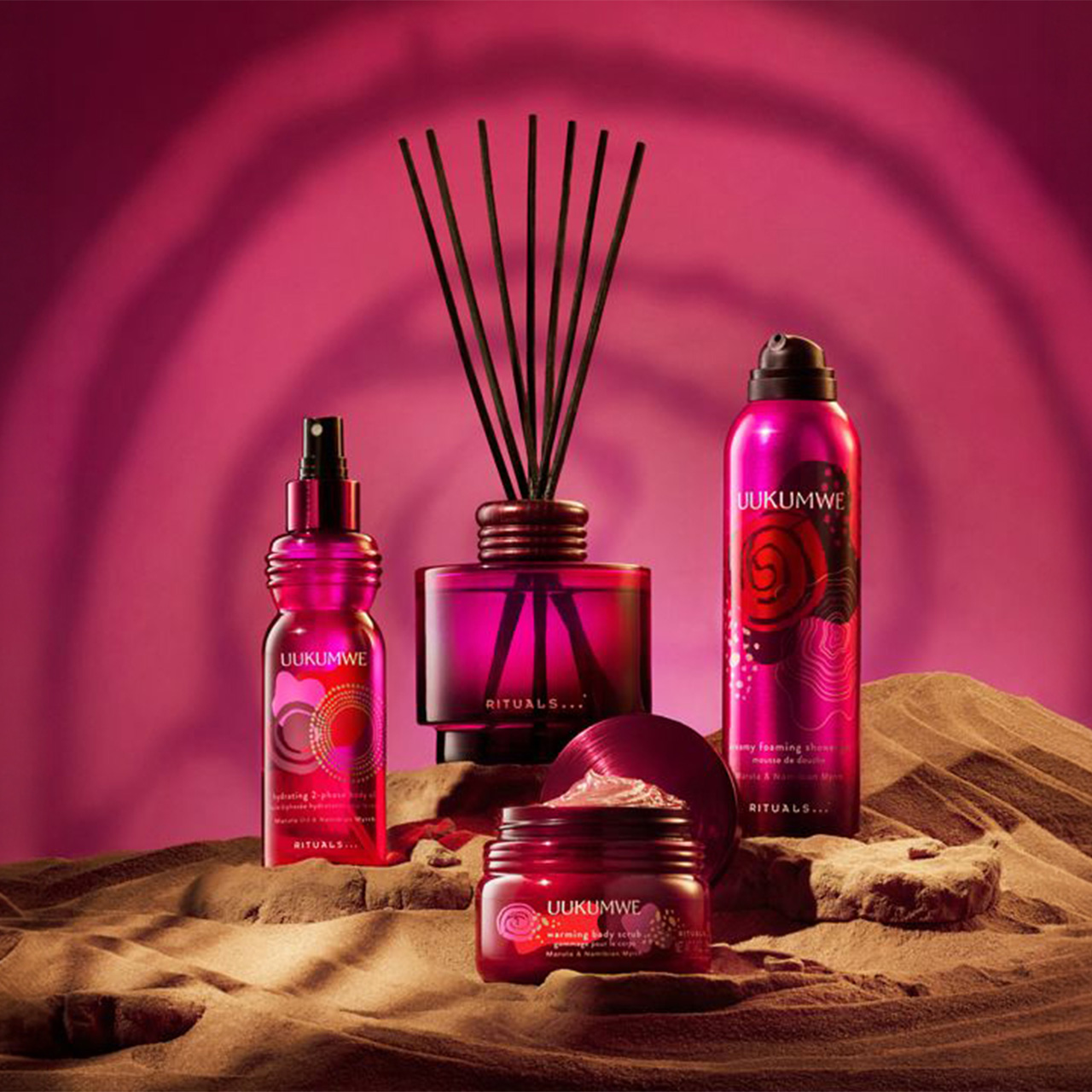 Pink-themed beauty products and a diffuser on a sandy surface with a vibrant, swirled background.
