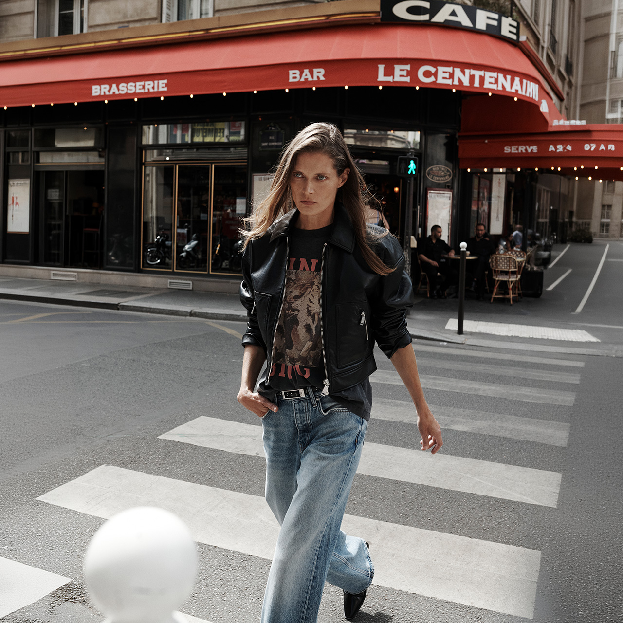 Woman wearing black leather jacket and graphic t-shirt with light blue jeans crossing street in front of Le Centenaire Parisian café