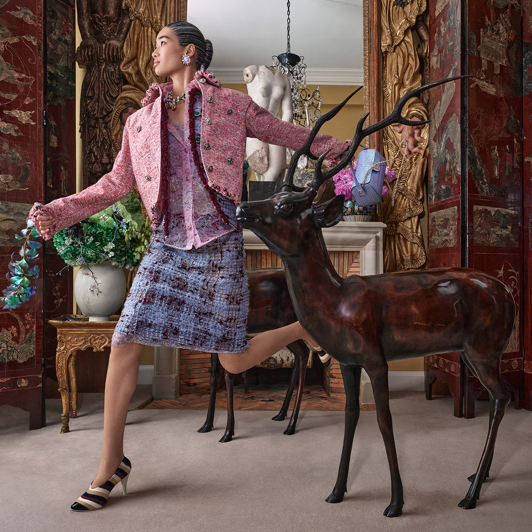 A woman poses in an opulent room, wearing a pink tweed jacket and a matching skirt from Chanel. She stands beside a decorative deer sculpture. The room features ornate wooden screens, a classic sculpture, and a vase with flowers, creating a luxurious atmosphere.
