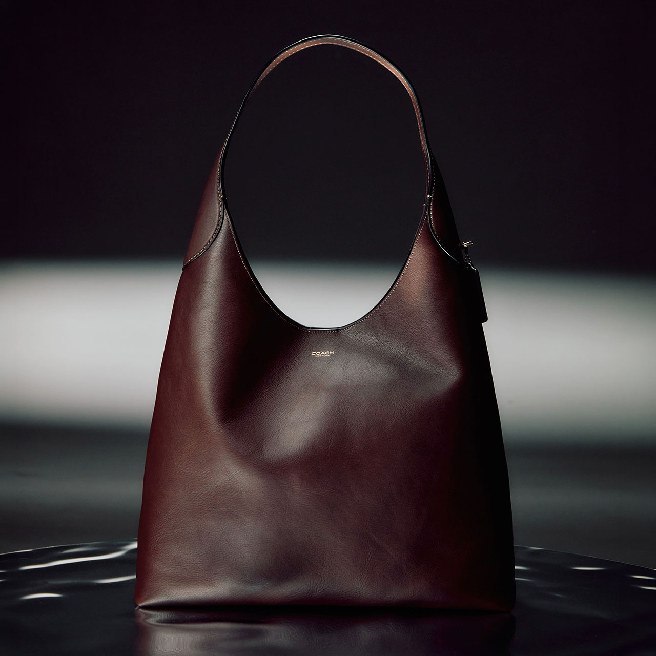 Deep burgundy leather shoulder bag with dual handles displayed against dark dramatic background with spotlight