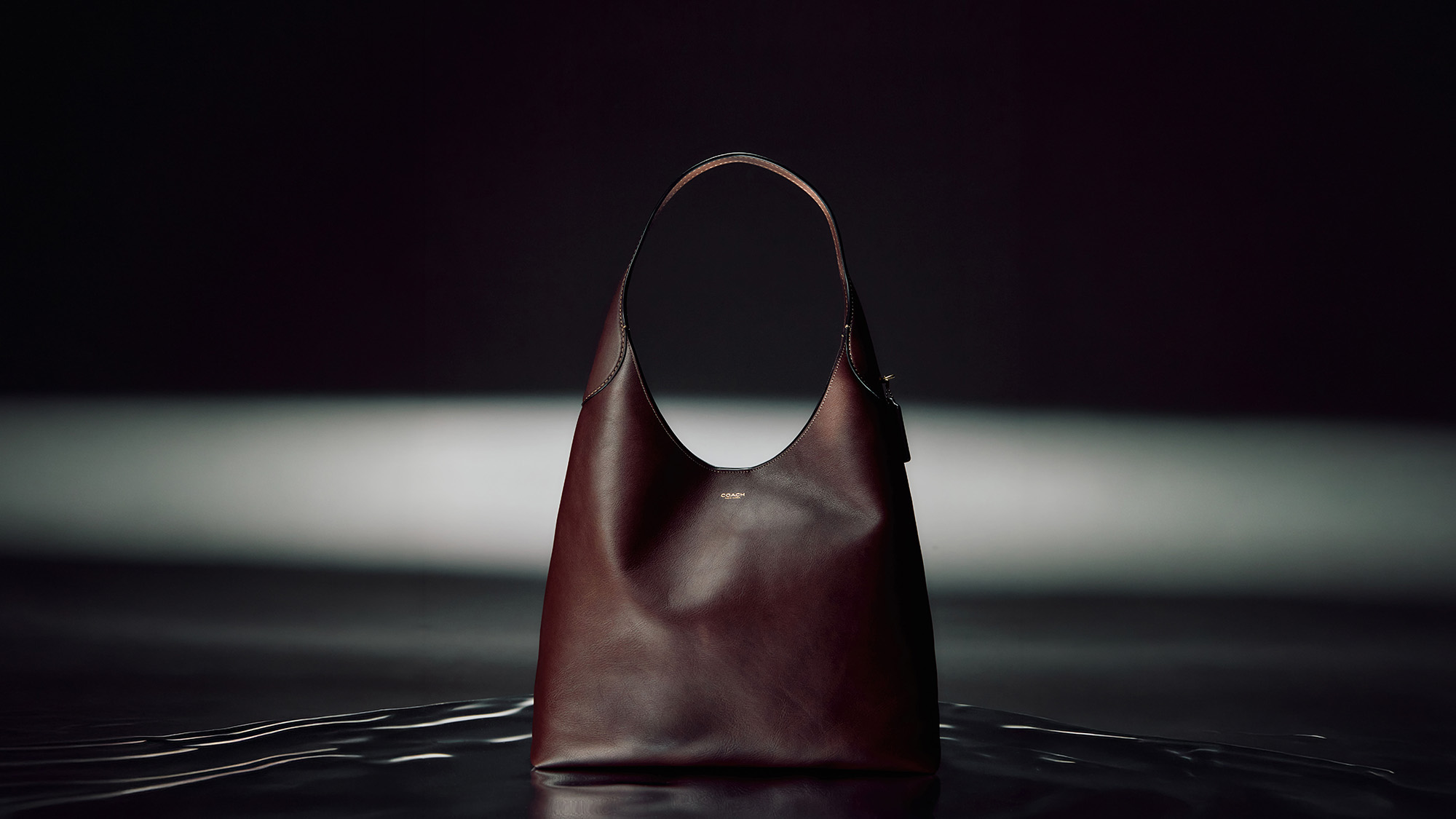 Deep burgundy leather shoulder bag with dual handles displayed against dark dramatic background with spotlight