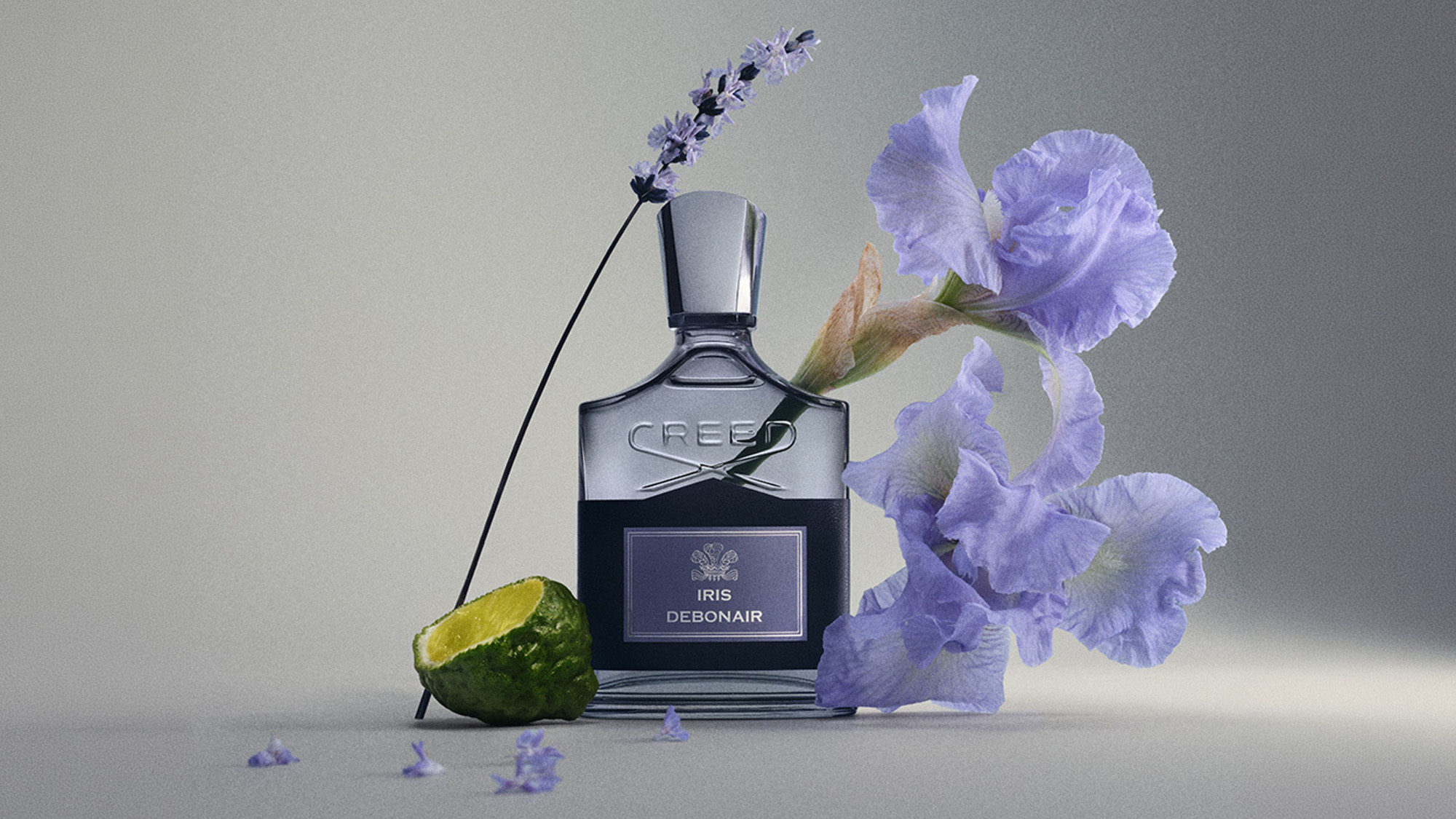 Creed Iris Debonair fragrance bottle surrounded by purple iris flowers and lime slice on neutral background