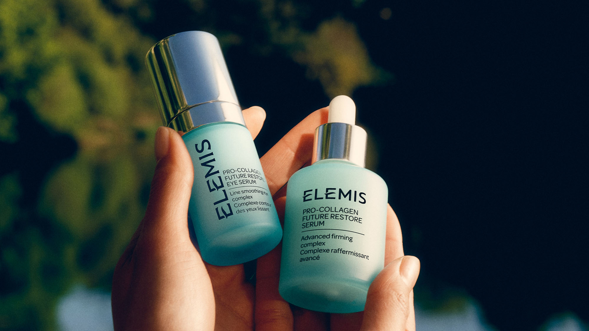 Two ELEMIS Pro-Collagen Future Restore serums in pale turquoise bottles held in hand against blurred green foliage background