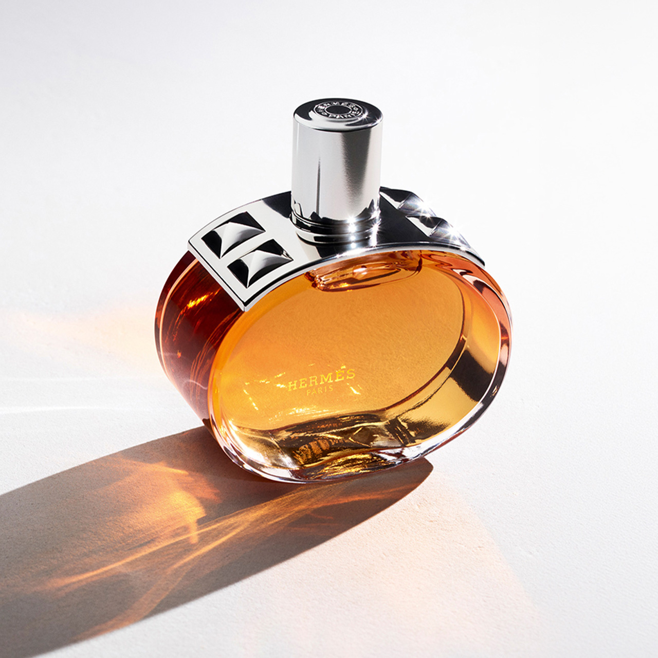 Hermès perfume bottle with amber liquid and silver-trimmed stopper casting golden shadow on white surface