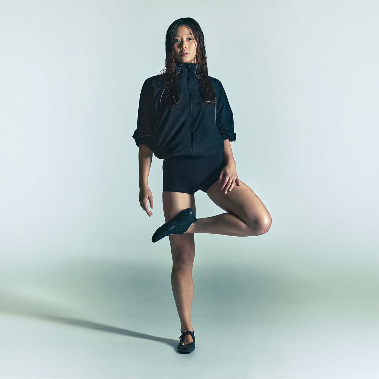 Woman wearing black zip-up jacket and black shorts with black trainers, posed against pale green background
