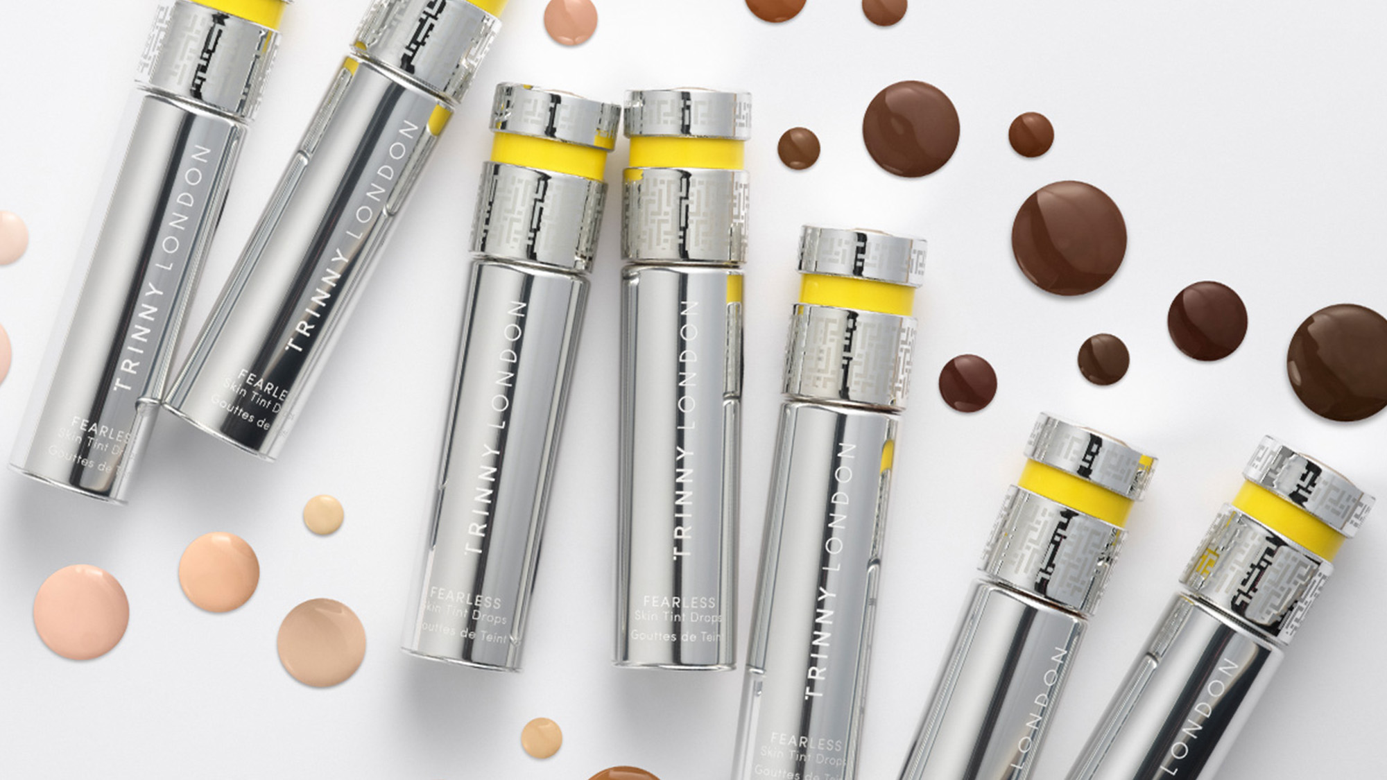 Selection of silver Trinny London makeup products with yellow accents and foundation swatches in various skin tones