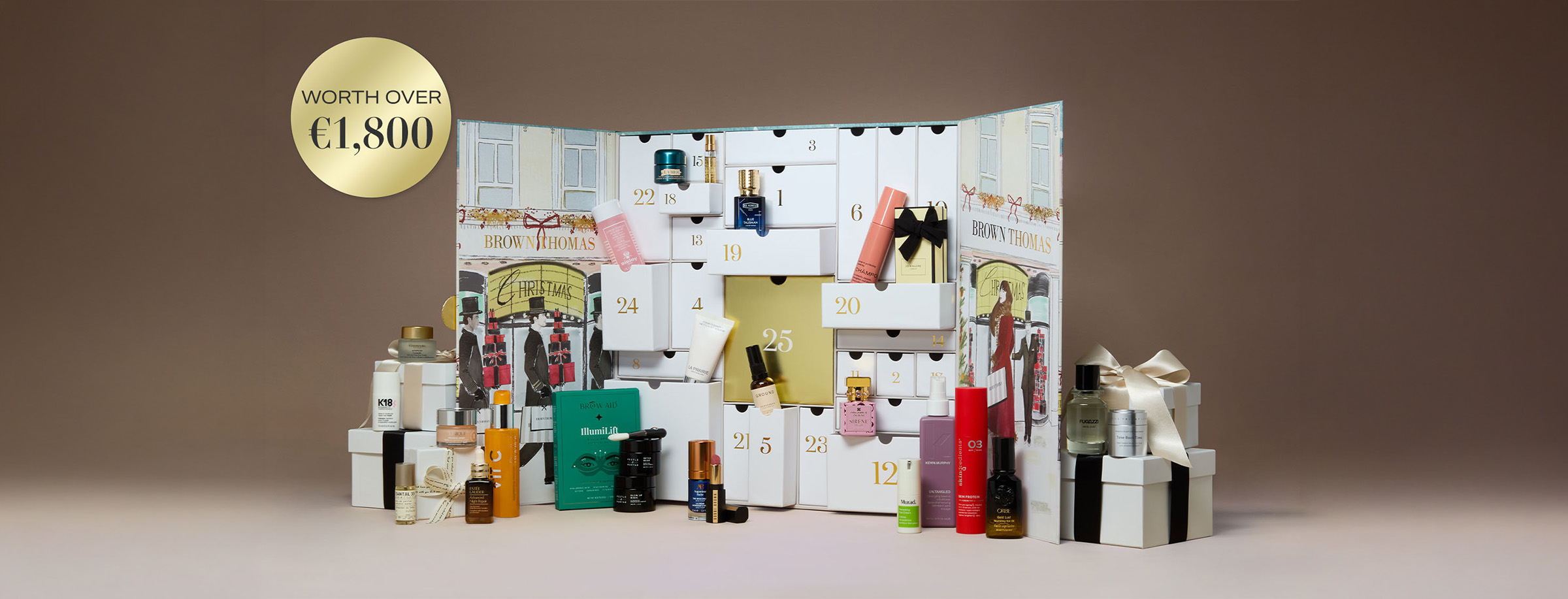 A luxurious advent calendar with multiple compartments filled with beauty products and small gifts is displayed. The calendar features festive illustrations and the Brown Thomas branding. A circular label in the corner reads 'Worth over €1,800.' Various skincare and cosmetic items are arranged in front.