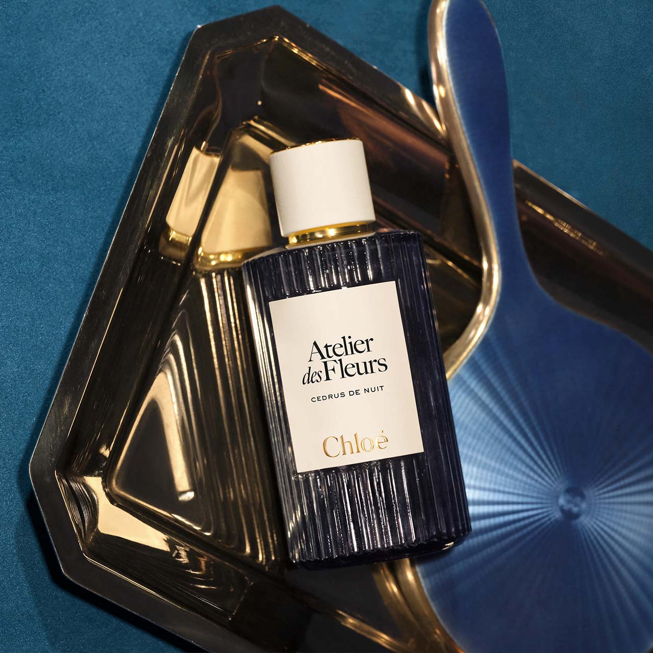 Chloé Atelier des Fleurs Cedrus de Nuit fragrance bottle in dark glass with white label, displayed on gold tray against blue background