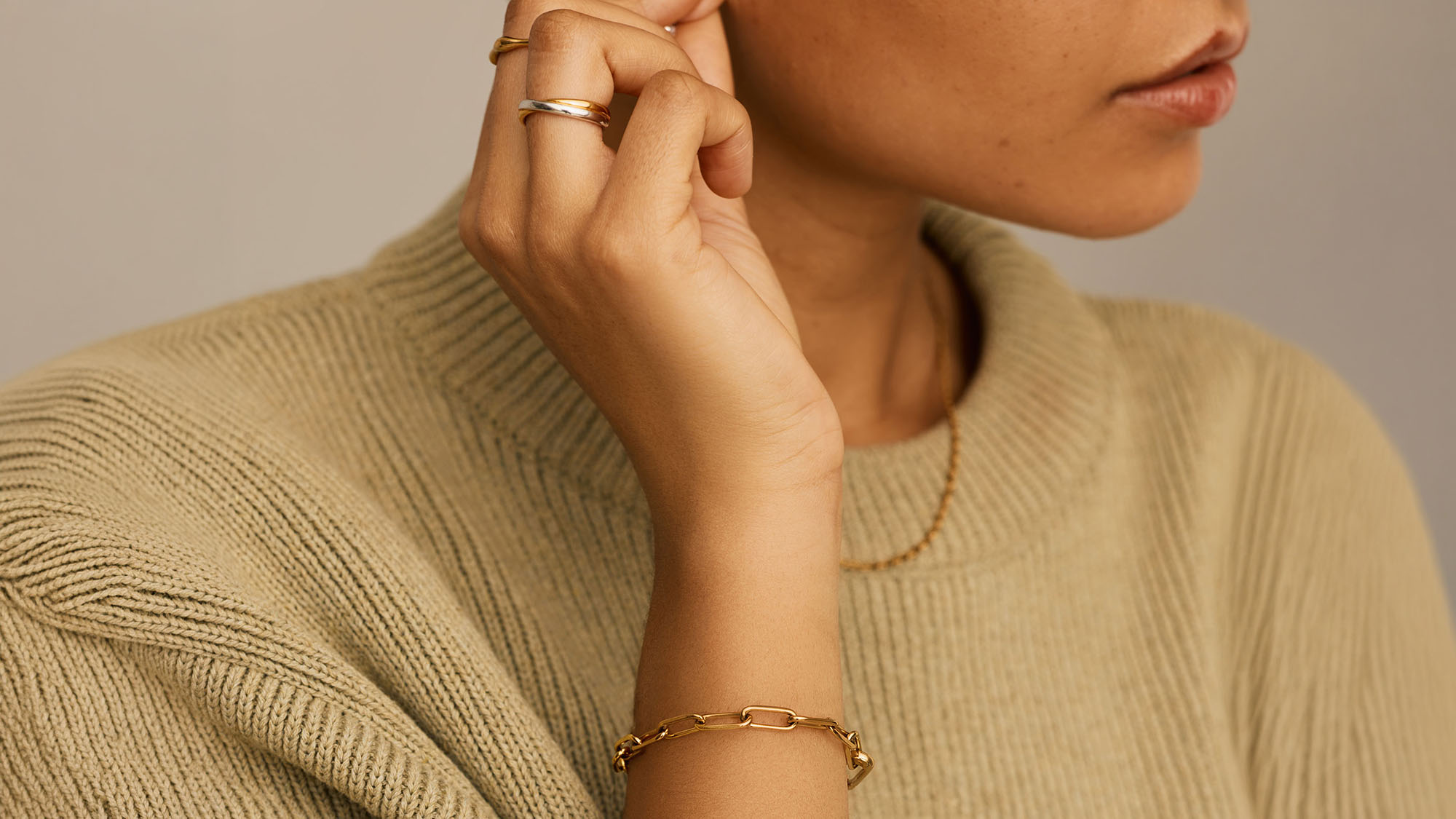 Woman wearing beige ribbed top with gold chain link bracelet and stacked gold rings on her hand