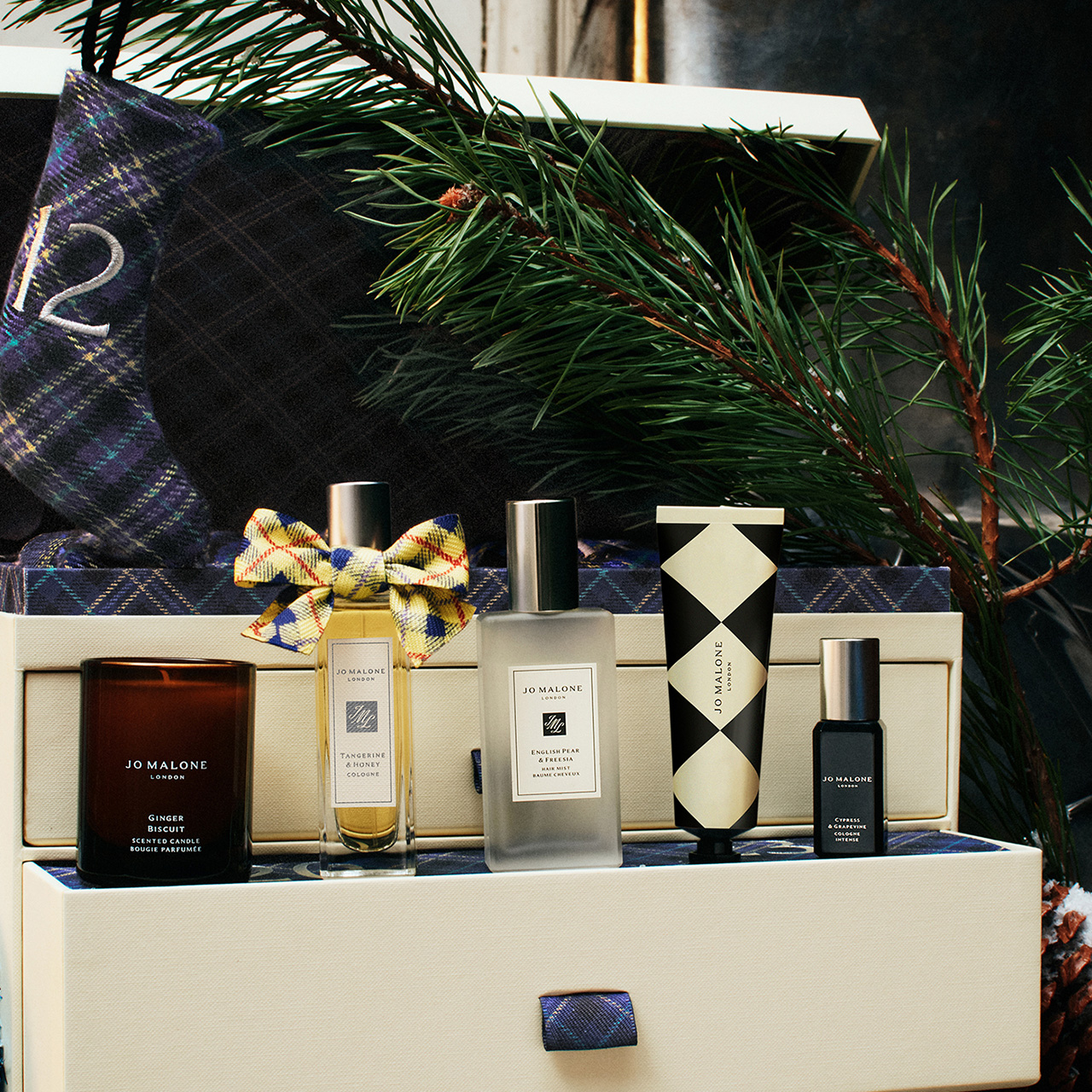 Jo Malone London beauty products including candles, colognes and a decorative box with tartan ribbon and festive greenery.