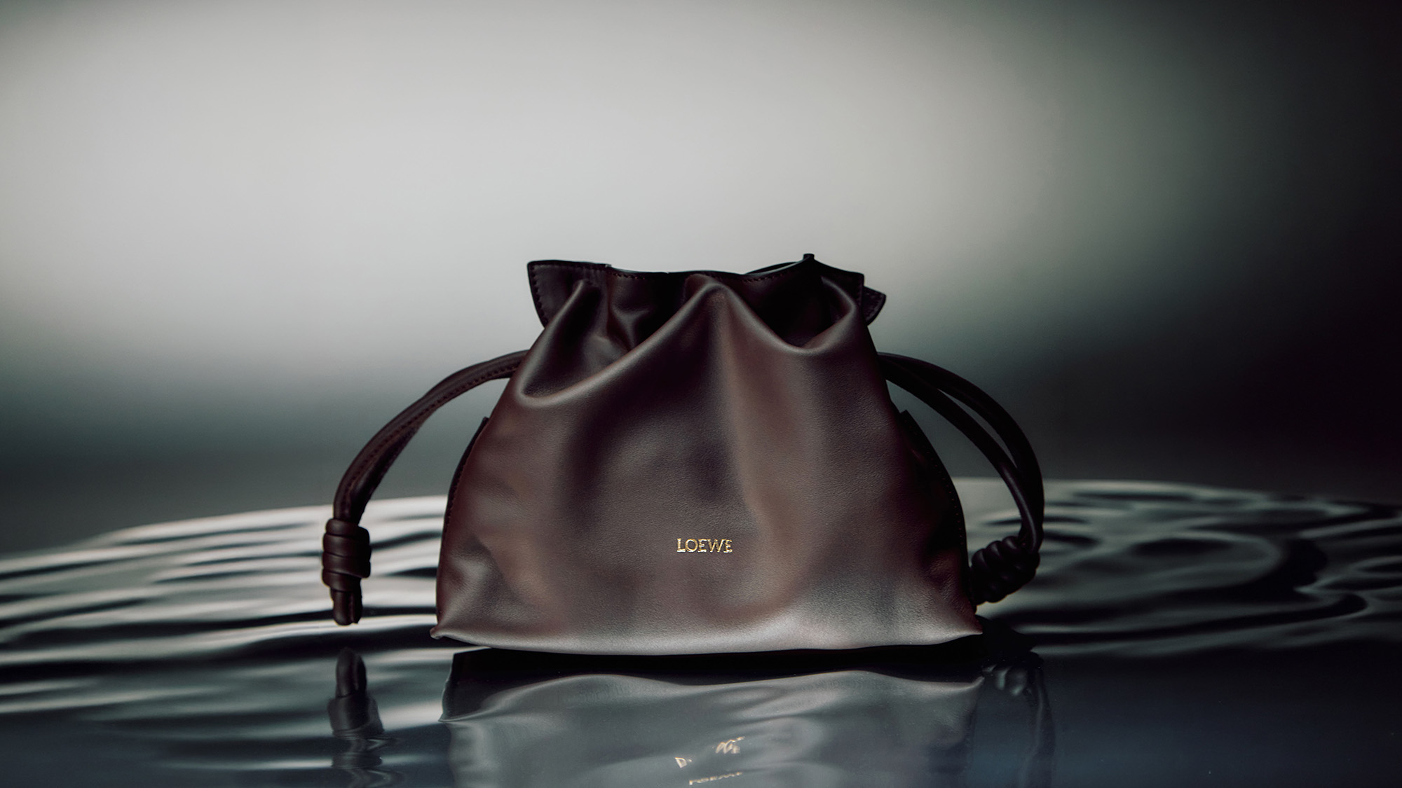 Dark burgundy leather bucket bag with drawstring handles and gold logo detail, displayed on reflective surface