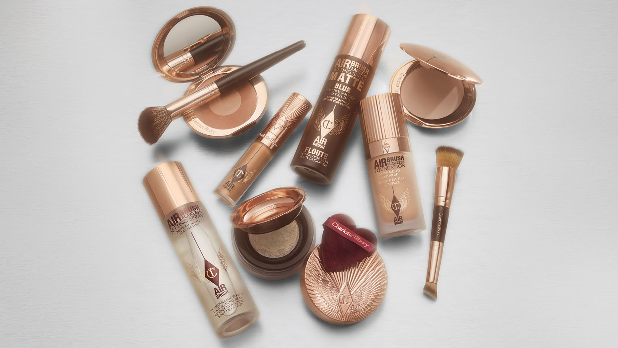 Collection of makeup products including foundations, powders, brushes, and blush in warm bronze and neutral tones arranged on white background