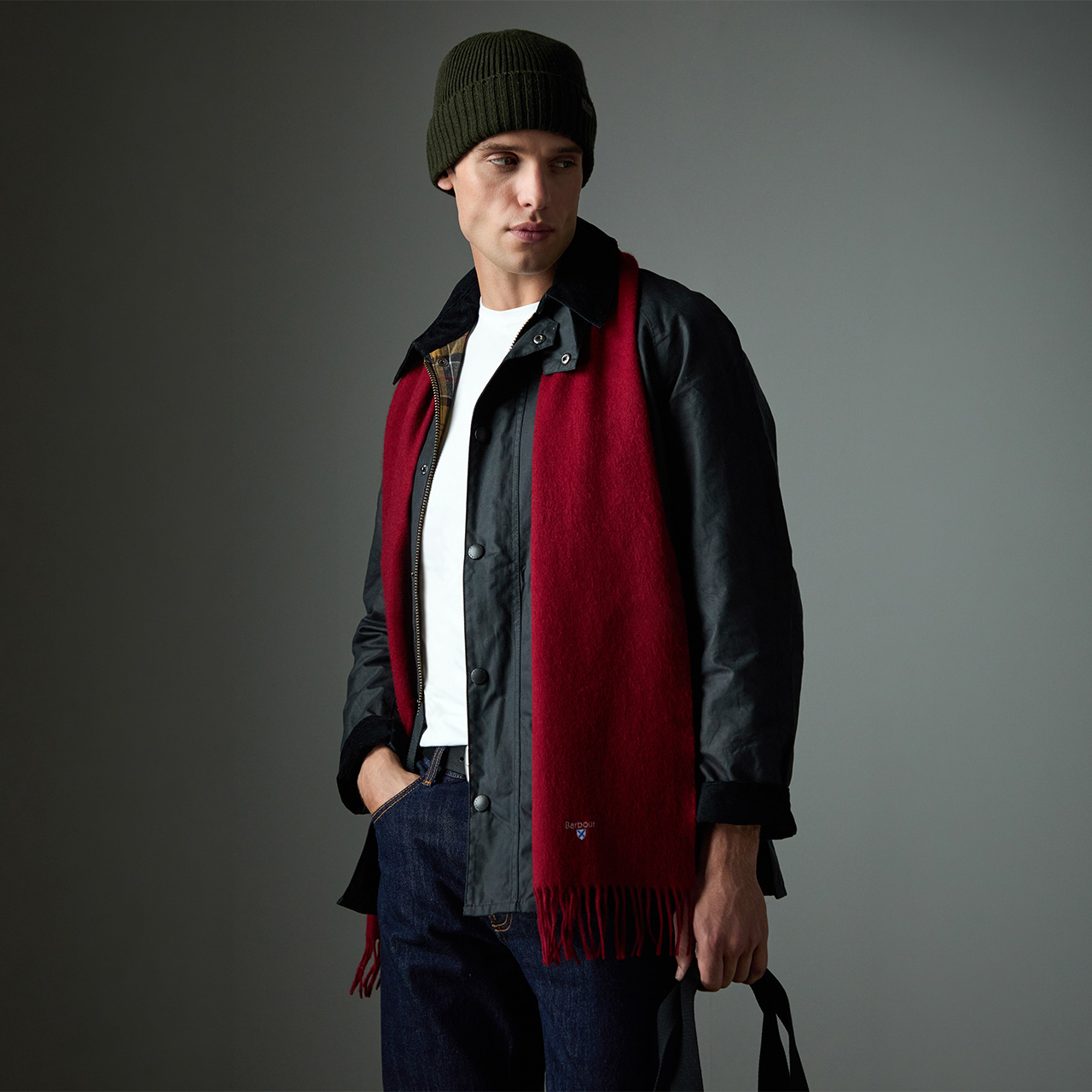 Man wearing dark jacket with red wool scarf, white t-shirt, dark beanie and dark jeans against neutral grey background.