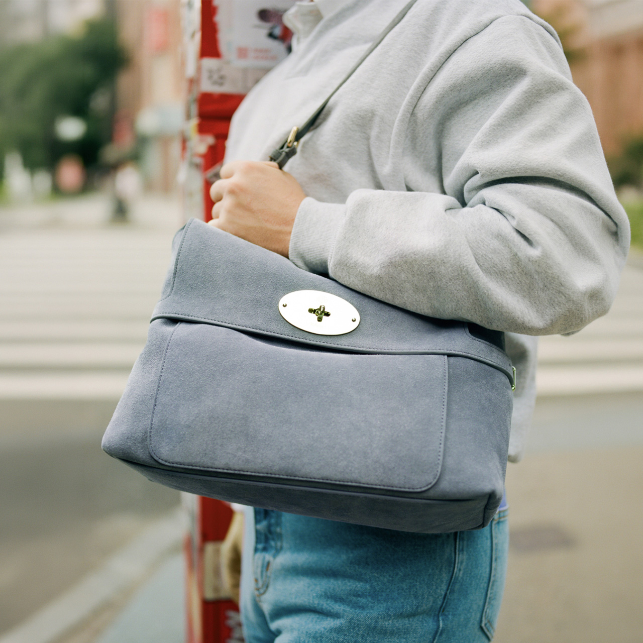 Person wearing grey hoodie carrying slate blue crossbody bag with oval gold hardware on urban street