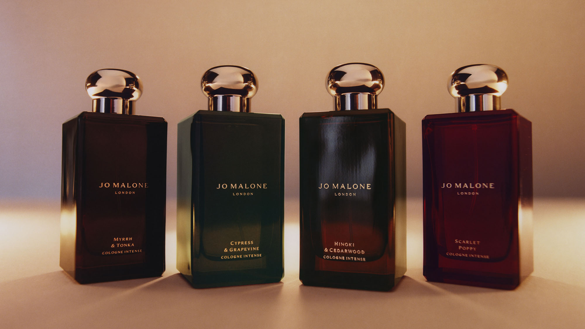 Four Jo Malone London cologne bottles in dark glass with gold caps, featuring scents Myrrh & Tonka, Cypress & Grapevine, Hinoki & Cedarwood, and Scarlet Poppy