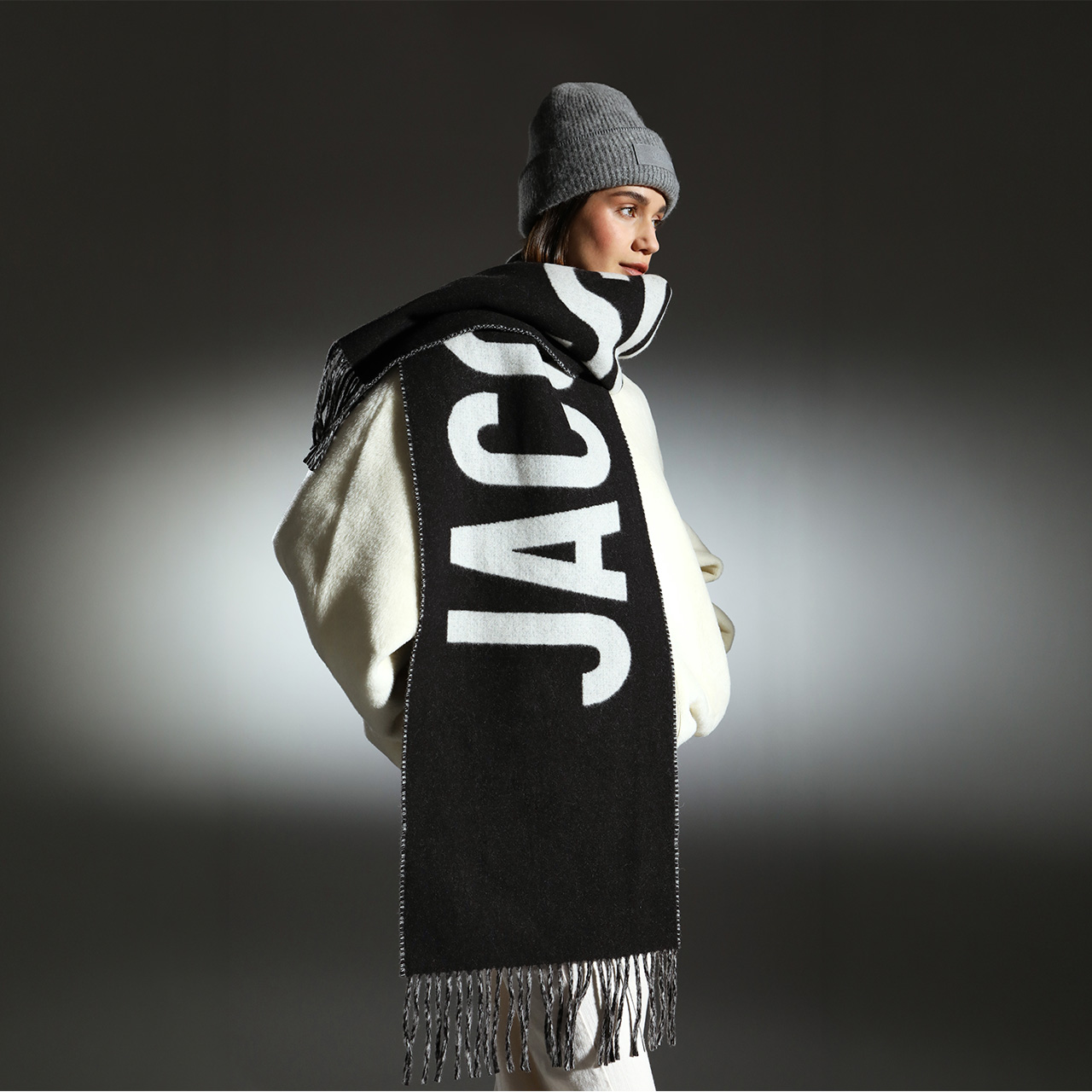 Woman wearing grey beanie and black and cream scarf with bold lettering, styled against neutral background
