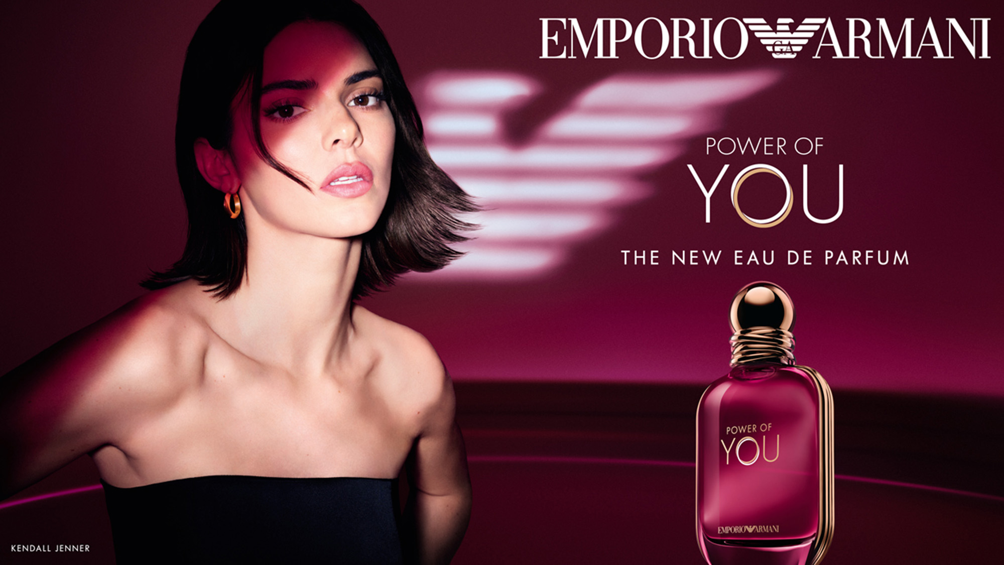 A woman poses beside a bottle of 'Emporio Armani Power of You' perfume against a maroon background.
