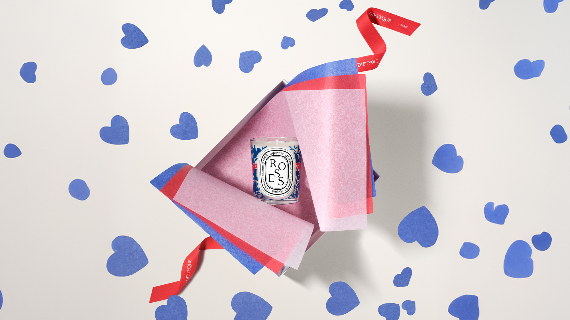 An open gift box with pink and red paper reveals a 'Roses' candle, surrounded by blue heart confetti.