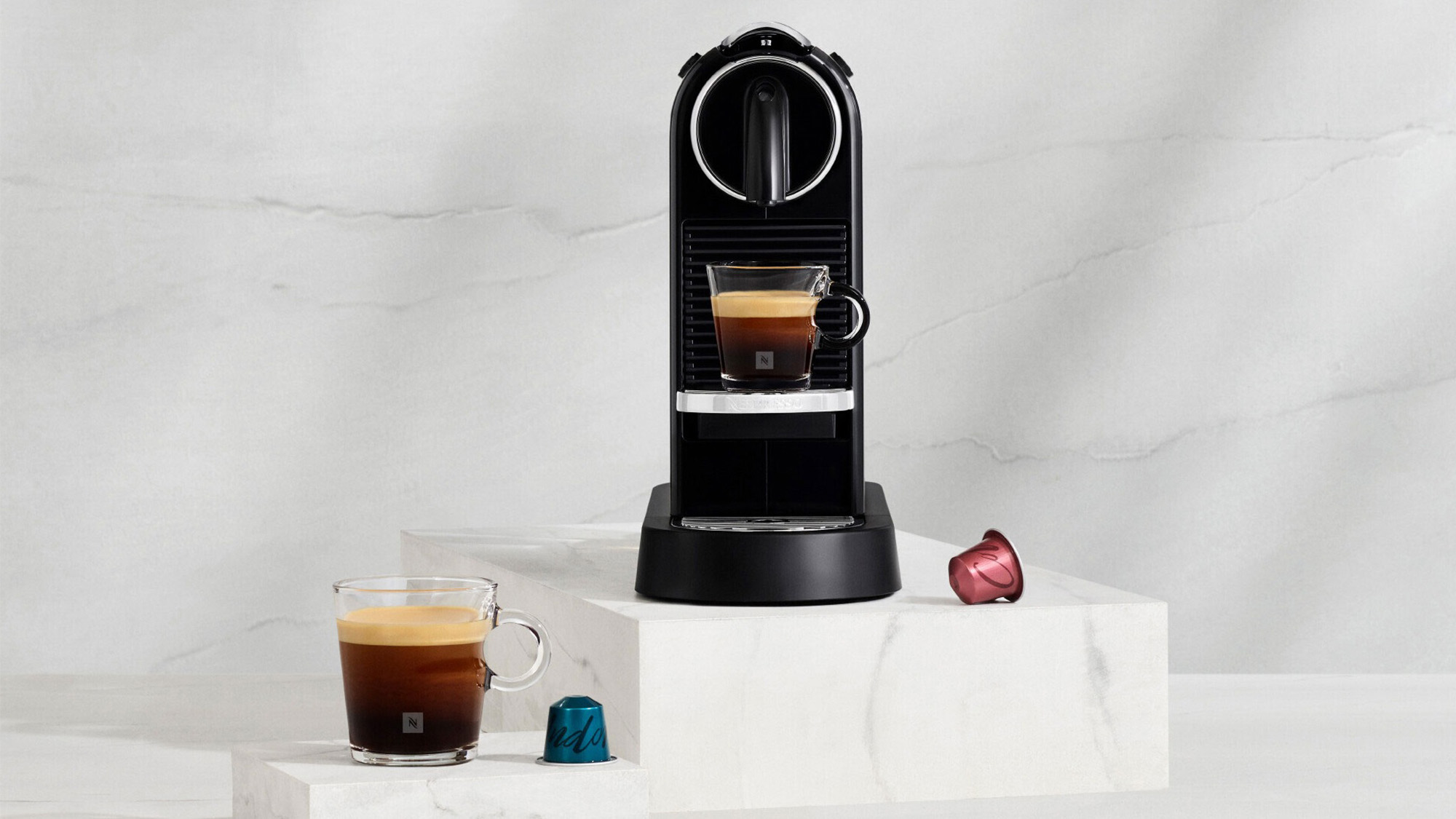 A black espresso machine on a marble surface with two coffee capsules and a cup of espresso nearby.