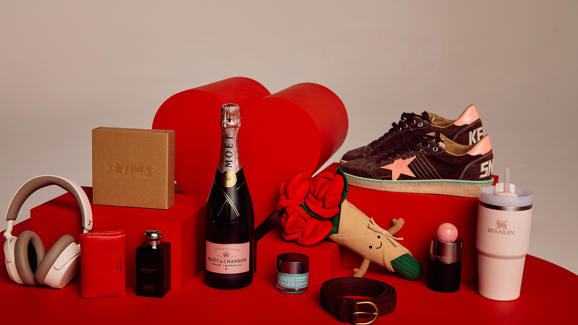 A collection of lifestyle items including headphones, a bottle of champagne, sneakers, skincare products, and a plush rose on a red surface.