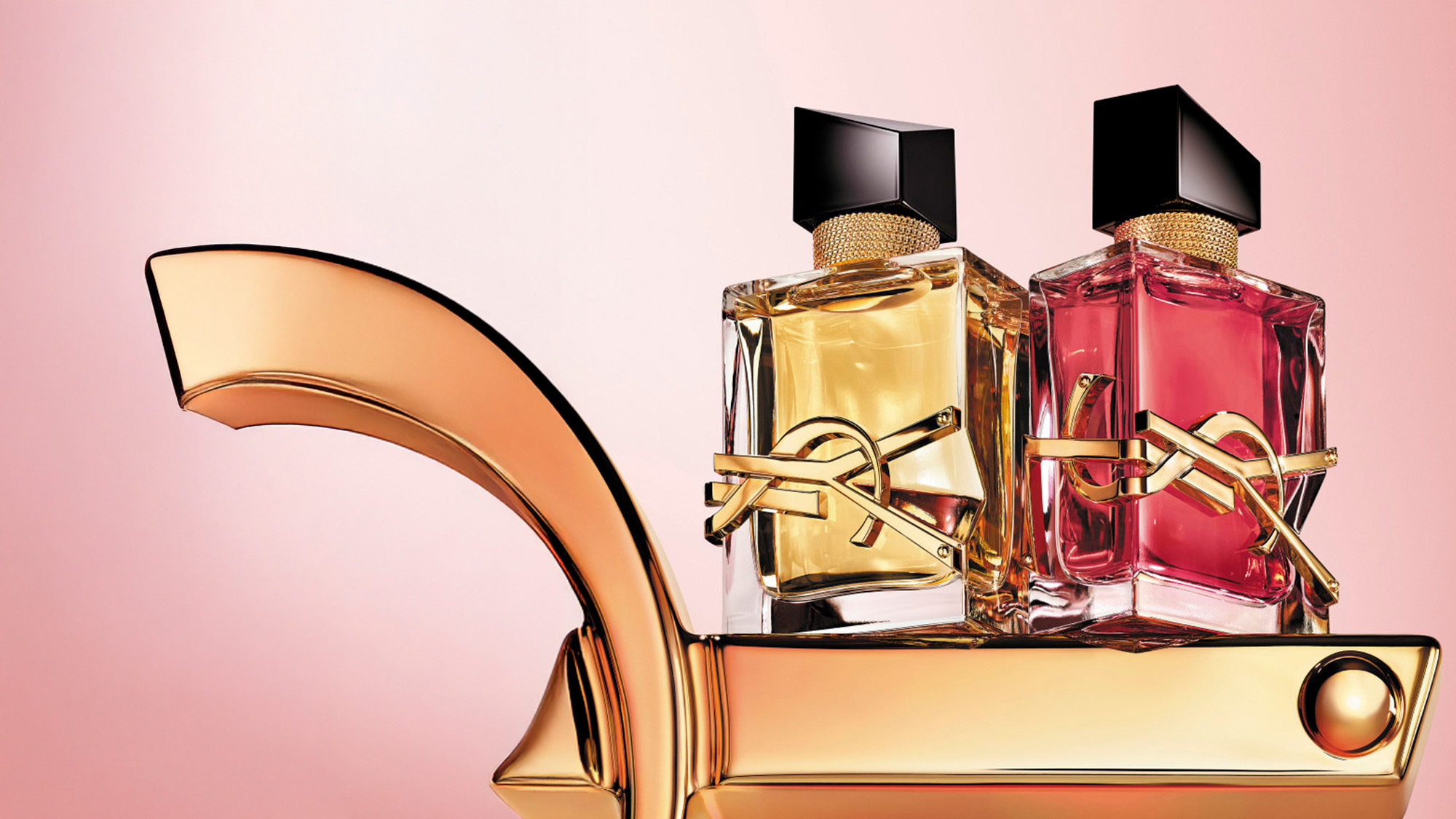 Two perfume bottles, one yellow and one pink, with black caps and gold accents, sit on a gold surface against a pink background.
