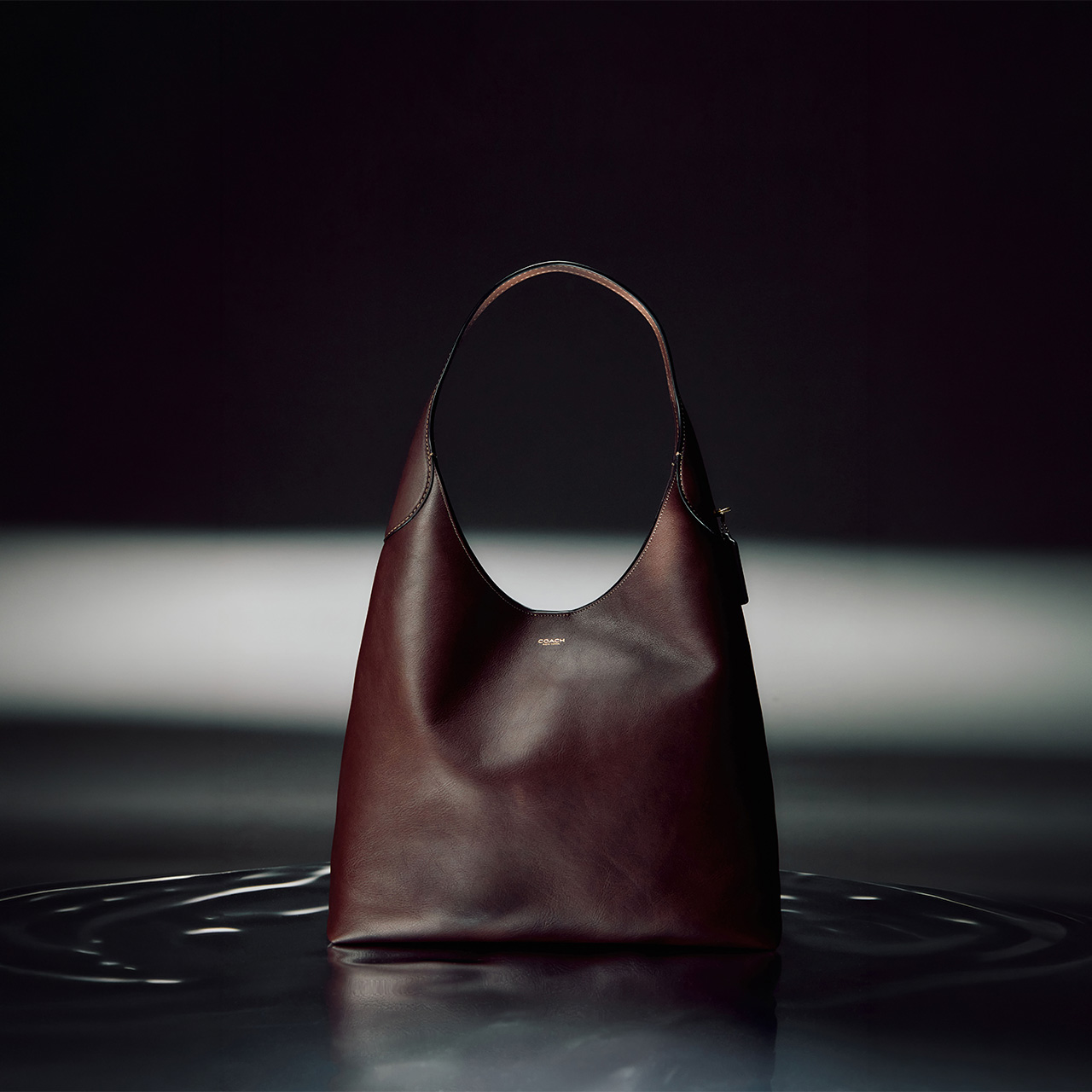 Burgundy leather hobo handbag with curved handles displayed against dramatic dark and light background