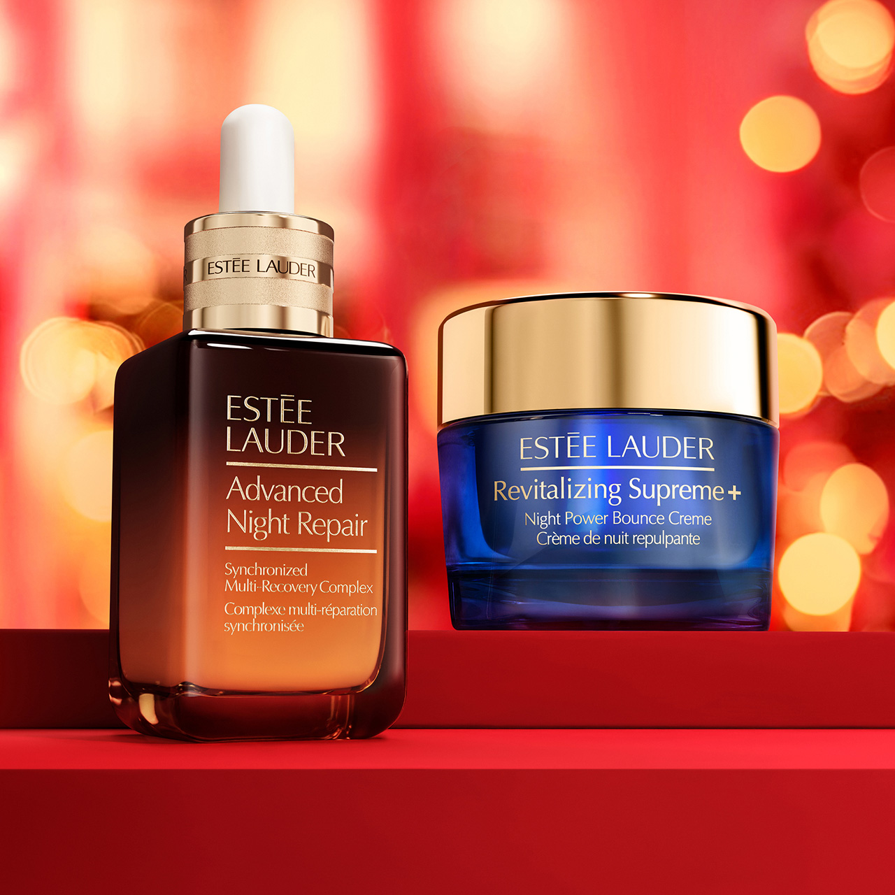 Estée Lauder Advanced Night Repair serum and Revitalising Supreme+ night cream displayed on red platform with festive golden bokeh background